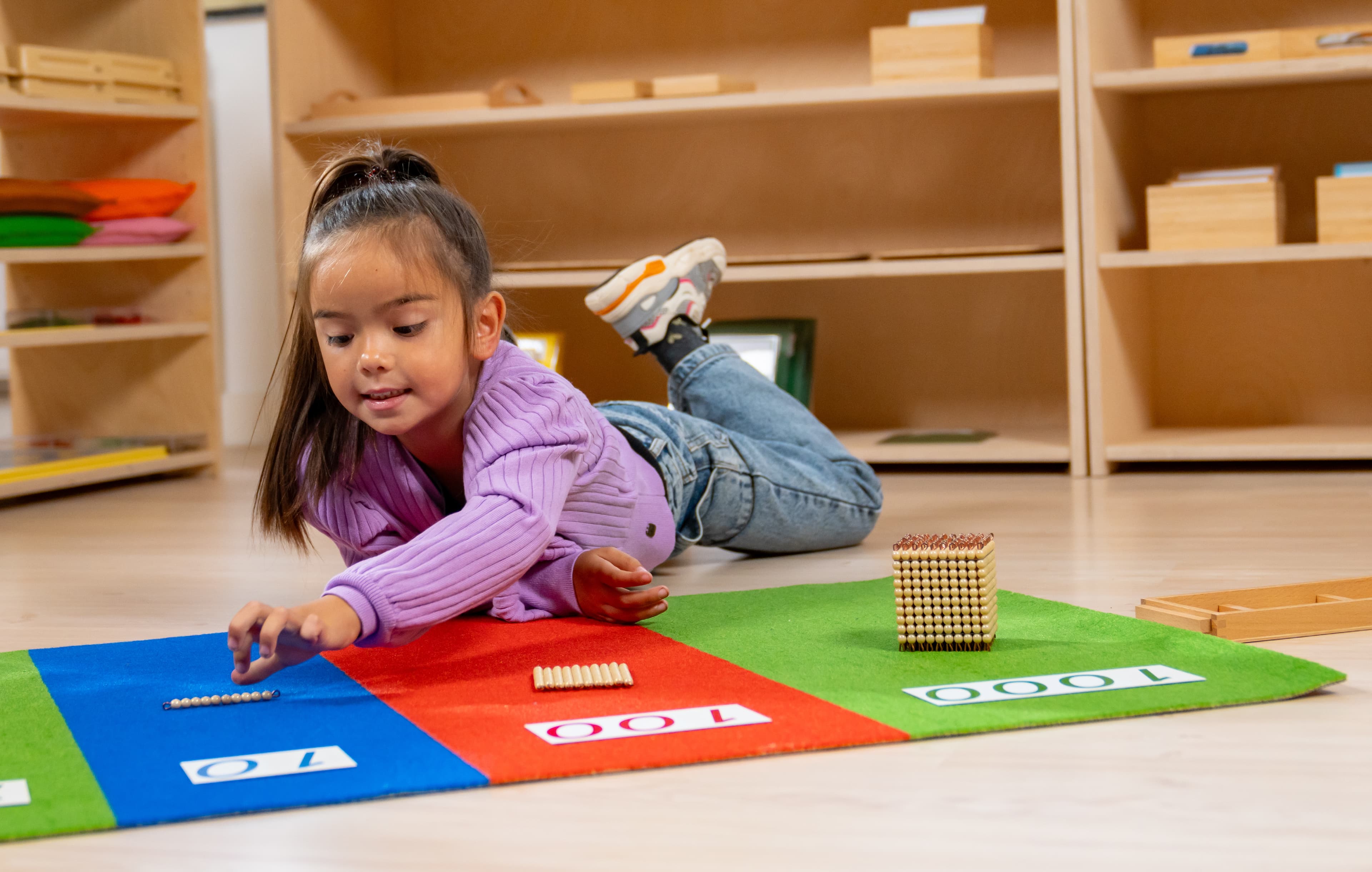 Ein Mädchen liegt auf dem Bauch mit einem Nienhuis Stellenwertteppich, auf dem sie Glasperlen ablegt. Im Hintergrund stehen Montessori-Schränke.