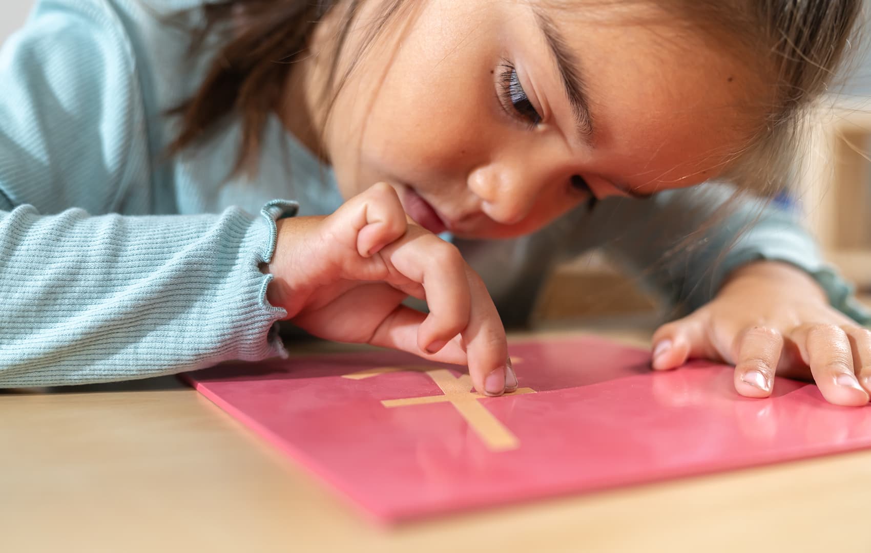 A girl traces a sandpaper letter on a pink board with her finger, concentrating on learning to write.